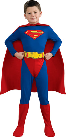 Boy posing in the DC Universe Classic Superman Costume featuring a full‑body long‑sleeve jumpsuit in bold blue with bright red lower accents, designed with a clean, streamlined silhouette that showcases Superman’s iconic look; the chest is printed with the unmistakable red‑and‑yellow “S” shield emblem, while a bright yellow printed belt adds contrast at the waist. The costume includes a flowing red cape, completing the classic superhero profile from head to toe