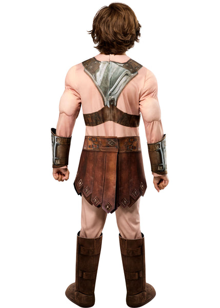 Back of boy in the Boys Deluxe Masters Of The Universe He-Man Costume  Featuring a full costume set including a flesh-toned jumpsuit with built-in muscle padding on the chest, arms, and legs, a molded chest armor piece with metallic detailing and a red cross emblem at the center, a brown faux leather-style loincloth skirt with stitched panel accents, matching wrist gauntlets, a wide belt with buckle detail, and tall boot tops that complete the armored warrior look.