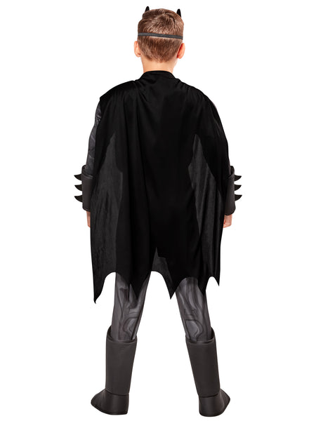 Boy modeling cape view of the DC Universe Deluxe Batman Costume featuring a full‑body gray and black superhero jumpsuit with built‑in padded muscle, Batman’s yellow‑and‑black bat emblem, a printed yellow utility belt graphic, black boot tops, a flowing black cape with scalloped edges, and a molded black Batman mask with pointed ears
