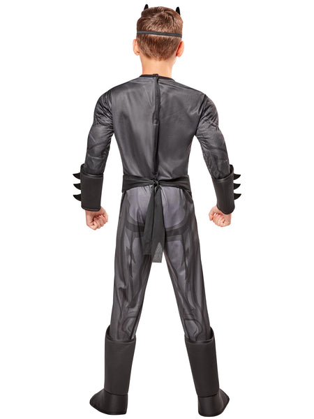 Boy showing the back of the DC Universe Deluxe Batman Costume featuring a full‑body gray and black superhero jumpsuit with built‑in padded muscle, Batman’s yellow‑and‑black bat emblem, a printed yellow utility belt graphic, black boot tops, a flowing black cape with scalloped edges, and a molded black Batman mask with pointed ears