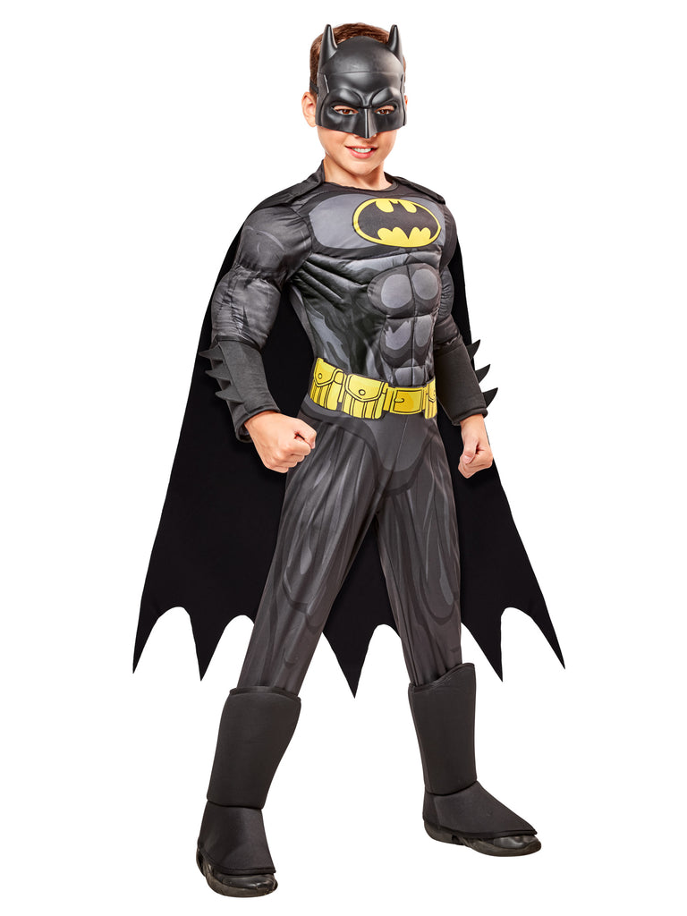 Boy superhero posing in the DC Universe Deluxe Batman Costume featuring a full‑body gray and black superhero jumpsuit with built‑in padded muscle, Batman’s yellow‑and‑black bat emblem, a printed yellow utility belt graphic, black boot tops, a flowing black cape with scalloped edges, and a molded black Batman mask with pointed ears