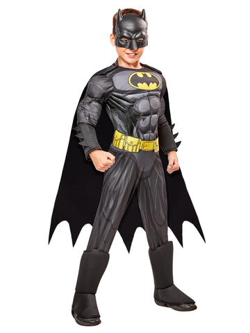 Boy superhero posing in the DC Universe Deluxe Batman Costume featuring a full‑body gray and black superhero jumpsuit with built‑in padded muscle, Batman’s yellow‑and‑black bat emblem, a printed yellow utility belt graphic, black boot tops, a flowing black cape with scalloped edges, and a molded black Batman mask with pointed ears