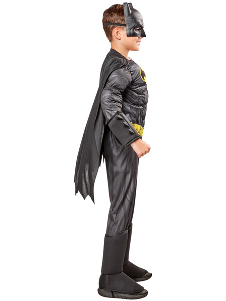 Boy showing side view of the DC Universe Deluxe Batman Costume featuring a full‑body gray and black superhero jumpsuit with built‑in padded muscle, Batman’s yellow‑and‑black bat emblem, a printed yellow utility belt graphic, black boot tops, a flowing black cape with scalloped edges, and a molded black Batman mask with pointed ears