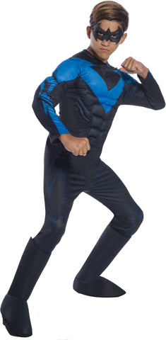 Boy in superhero pose wearing the Deluxe Nightwing Costume featuring a sleek, full‑body black jumpsuit with built‑in padded muscle detailing across the chest, arms, and legs to create a powerful superhero silhouette; the suit is highlighted with Nightwing’s signature bold blue wing‑shaped emblem stretching across the chest and shoulders, with matching blue accent graphics running along the arms and legs. Also includes tall integrated boot tops that extend up the calves and a black domino‑style eye mask