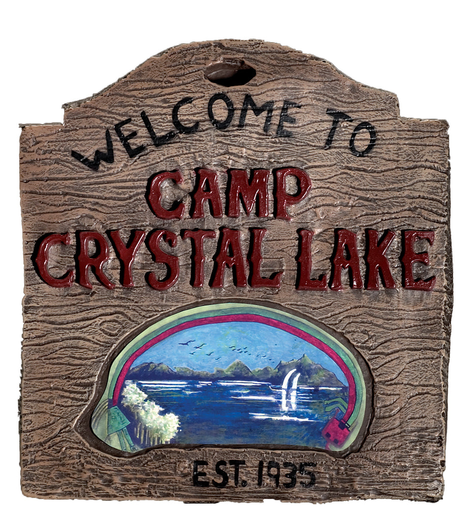 A lightweight vacuform plastic sign molded to resemble aged, weathered wood, designed in the iconic camp sign shape with a rounded top and notched edges; detailed with raised faux wood‑grain texture throughout and bold painted lettering that reads “WELCOME TO CAMP CRYSTAL LAKE” across the front, with “EST. 1935” stamped near the bottom; centered with a painted lake scene vignette showing dark blue water, distant trees, and shoreline details that evoke the eerie setting from the legendary horror franchise