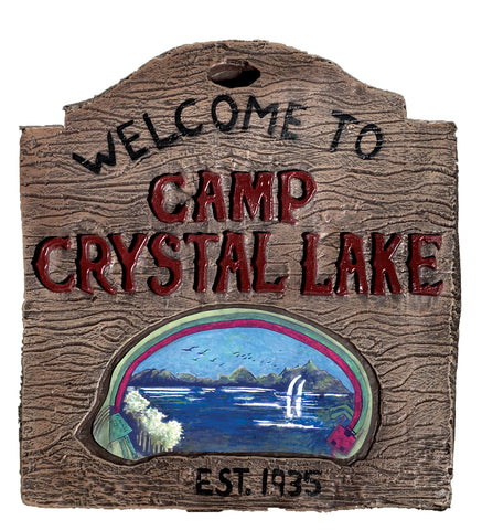 A lightweight vacuform plastic sign molded to resemble aged, weathered wood, designed in the iconic camp sign shape with a rounded top and notched edges; detailed with raised faux wood‑grain texture throughout and bold painted lettering that reads “WELCOME TO CAMP CRYSTAL LAKE” across the front, with “EST. 1935” stamped near the bottom; centered with a painted lake scene vignette showing dark blue water, distant trees, and shoreline details that evoke the eerie setting from the legendary horror franchise