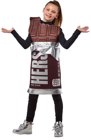 Girl posing in Kids Hershey's Chocolate Bar Costume featuring an oversized tunic designed to resemble a classic Hershey’s milk chocolate bar, printed in rich brown tones with highly detailed “chocolate square” segments molded into the top portion and stamped with the HERSHEY’S logo; the lower half displays the iconic brown wrapper design with bold white “HERSHEY’S” lettering, “Since 1894” text, and a silver foil‑style trim printed around the waist and hem, creating a realistic candy‑bar wrapper illusion