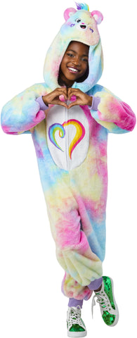 Girl posing Togetherness Bear Costume featuring soft, plush one‑piece jumpsuit covered in a pastel rainbow tie‑dye pattern blending pink, blue, yellow, and purple tones; a cozy hood shaped like the Togetherness Bear, complete with rounded bear ears, an embroidered smiling face, and small heart details on the cheeks. Front of the costume includes a white belly panel printed with the signature rainbow heart Care Bears symbol, while the back is finished with an attached fluffy pink bear tail and a small heart 