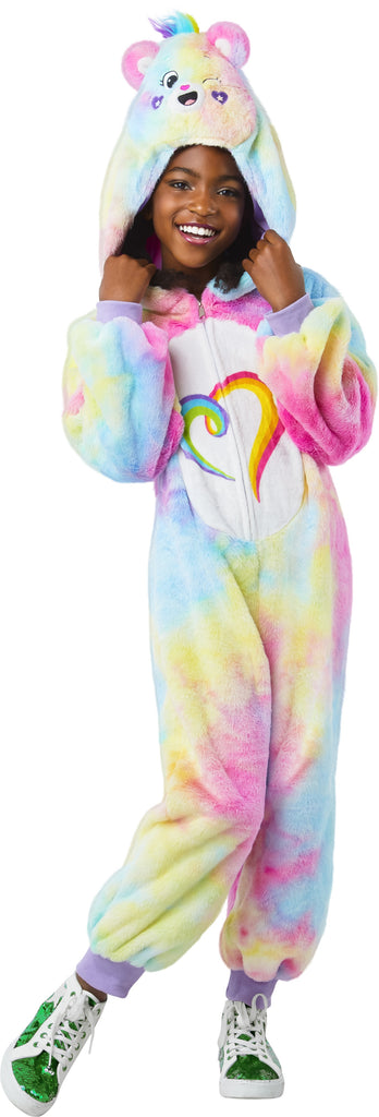 Girl posing Togetherness Bear Costume featuring soft, plush one‑piece jumpsuit covered in a pastel rainbow tie‑dye pattern blending pink, blue, yellow, and purple tones; a cozy hood shaped like the Togetherness Bear, complete with rounded bear ears, an embroidered smiling face, and small heart details on the cheeks. Front of the costume includes a white belly panel printed with the signature rainbow heart Care Bears symbol, while the back is finished with an attached fluffy pink bear tail and a small heart 