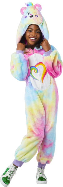 Girl posing Togetherness Bear Costume featuring soft, plush one‑piece jumpsuit covered in a pastel rainbow tie‑dye pattern blending pink, blue, yellow, and purple tones; a cozy hood shaped like the Togetherness Bear, complete with rounded bear ears, an embroidered smiling face, and small heart details on the cheeks. Front of the costume includes a white belly panel printed with the signature rainbow heart Care Bears symbol, while the back is finished with an attached fluffy pink bear tail and a small heart 