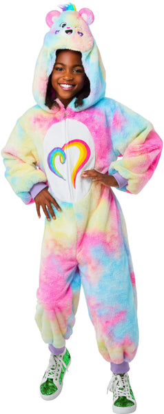 Girl posing Togetherness Bear Costume featuring soft, plush one‑piece jumpsuit covered in a pastel rainbow tie‑dye pattern blending pink, blue, yellow, and purple tones; a cozy hood shaped like the Togetherness Bear, complete with rounded bear ears, an embroidered smiling face, and small heart details on the cheeks. Front of the costume includes a white belly panel printed with the signature rainbow heart Care Bears symbol, while the back is finished with an attached fluffy pink bear tail and a small heart 