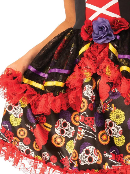 Close of the Girls Day Of The Dead Costume Dress featuring a multi‑layered black dress designed with an off‑the‑shoulder neckline trimmed in ruffled lace bands accented in purple, yellow, and gold tones; bodice with a red crisscross panel at the front and a decorative floral accent at the waist, full skirt  with colorful sugar skulls, flowers, butterflies, and festive motifs, finished with cascading red lace trim along the hem, black headpiece adorned with red fabric roses and a sheer black veil