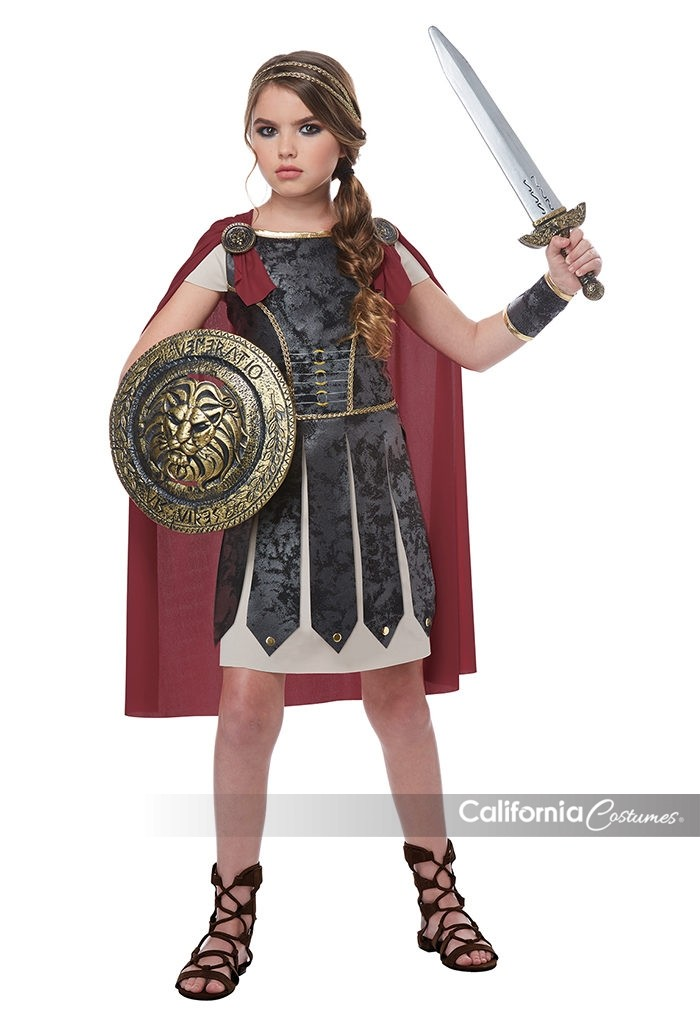 Girl posing in the Girls Fearless Roman or Greek Gladiator Costume Dress Featuring a dress with a black suede-look armor-style bodice, structured panel detailing with gold trim, attached short tan sleeves, and a tan skirt with layered pointed panels, along with a detachable red cape secured at the shoulders with two round medallion pins, a braided headband, and a single wide wrist cuff.
