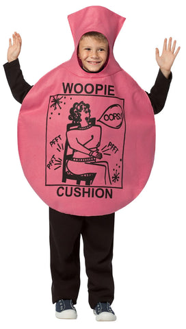 Child in the Kids Woopie Cushion Costume Featuring a one-piece oversized cushion-shaped tunic in pink polyester felt with a rounded, inflated silhouette and a tall nozzle-style top. The front displays black printed artwork including the word woopie above and cushion below, along with a cartoon-style illustration and small graphic details. The design includes a circular face opening at the top and wide side openings for arms