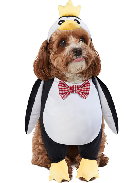 Cockapoo puppy modeling the Pets Walking Penguin Costume which features a plush, full‑body penguin "walking" illusion in classic black‑and‑white coloring with a soft white belly panel, black flipper‑style sides, and oversized yellow plush feet and an attached penguin character hood with embroidered eyes, a soft yellow beak, and a bright yellow crown perched on top, along with a red‑and‑white checkered bow tie