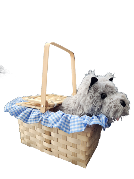 Side view of Boots and Brother's Puppy in a Basket featuring a woven natural wood basket with a handle, lined with a blue and white gingham fabric ruffle, holding a plush dog toy with gray and white fur, black ears, and a pink tongue