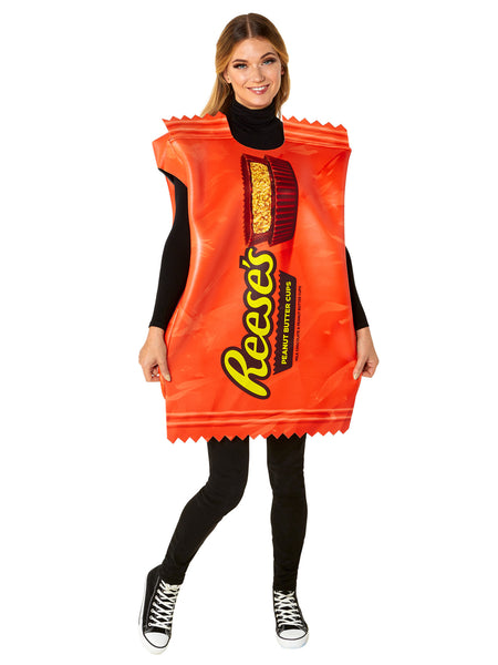 Woman posing in the Adults Reese's Peanut Butter Cup Candy Costume featuring a bold, oversized tunic designed to resemble a classic Reese’s wrapper, printed in vibrant orange with large yellow “Reese’s” lettering and a detailed image of a sliced peanut butter cup at the top; styled with crimped “wrapper” edges at the neckline and hem for a realistic candy‑bar effect. The lightweight, sleeveless design slips easily over clothing, creating a full‑body candy package silhouette that’s instantly recognizable 