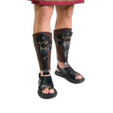 Featuring a matching pair of shin‑high armored leg coverings designed in textured brown faux‑leather, shaped to wrap securely around the lower legs from calf to ankle. Each guard is accented with layered black faux‑leather straps, round gold‑tone studs, and a prominent circular gold ring centerpiece at the front, creating a structured, battle‑ready look inspired by ancient Roman armor. The guards taper cleanly at the ankle with strap extensions that sit just above the foot